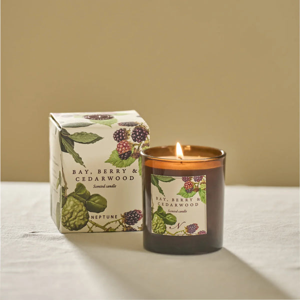 A lit amber glass scented candle stands next to its illustrated product box on a light fabric surface against a warm beige background. The label and box read Bay, Berry & Cedarwood and Neptune.