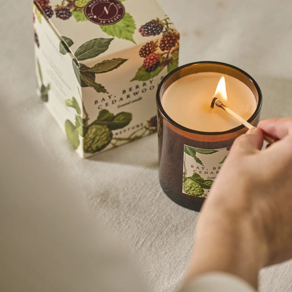 A lit amber glass jar candle with a visible flame, shown next to its illustrated product box and a hand holding a match. The label reads Bay Berry Cedarwood.
