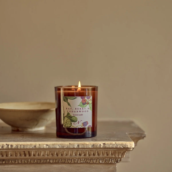 A lit amber glass candle jar with a botanical label reading 'Bay, Berly & Cedarwood' sits on a stone ledge against a warm beige wall.