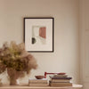 A framed abstract art print with minimalist shapes in muted beige, terracotta, and dark olive tones, hanging on a warm cream wall above a wooden console table styled with books and a vase of dried grass.