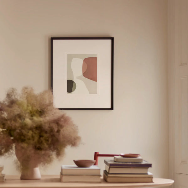 A framed abstract art print with minimalist shapes in muted beige, terracotta, and dark olive tones, hanging on a warm cream wall above a wooden console table styled with books and a vase of dried grass.