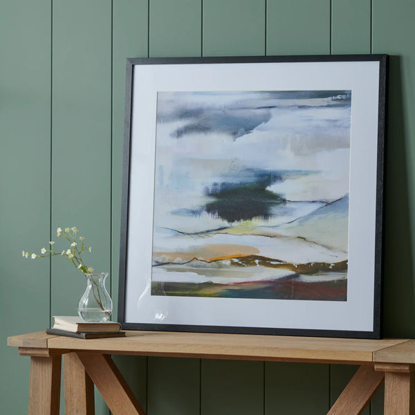 A framed abstract landscape painting with a wide white mat and black frame, leaning against a dark green paneled wall on a wooden table next to a small vase with white flowers.
