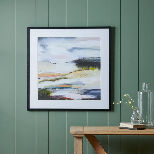 A framed abstract landscape painting with a white mat and black frame, hanging on a green paneled wall above a wooden table with books and a glass vase.