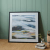 A framed abstract landscape painting with a white mat and black frame, hanging on a green paneled wall above a wooden table with books and a glass vase.