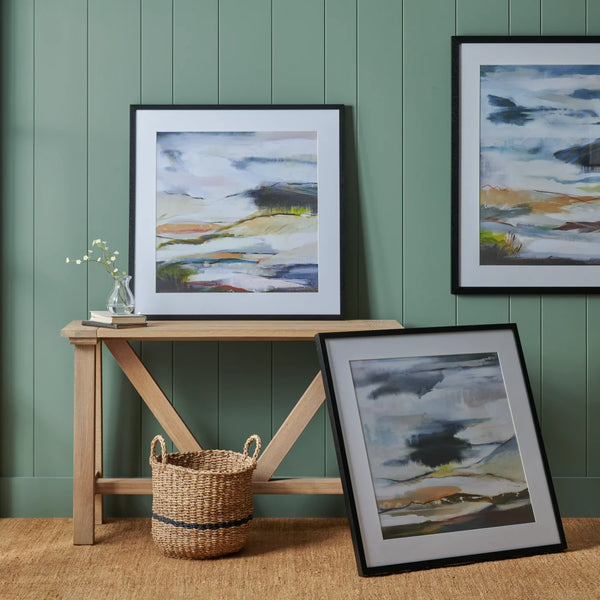 Three framed abstract landscape paintings are displayed in a styled room setting with a green paneled wall, a light wood console table, a woven basket, and a small vase with white flowers.