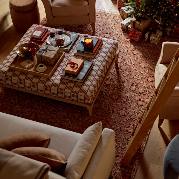 A styled living room scene with a patterned ottoman coffee table in the center, surrounded by sofas, a Christmas tree with wrapped gifts, and warm ambient lighting.