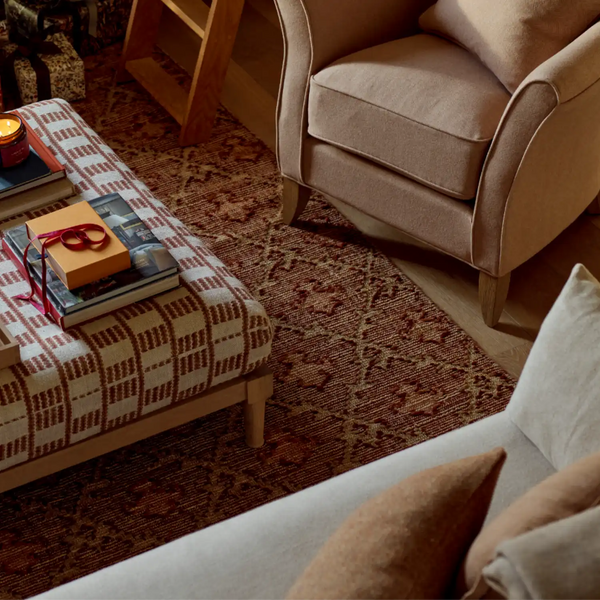 A cozy living room scene showing a patterned ottoman, a beige armchair, a light sofa, and a rust-red patterned area rug with wooden furniture and books.