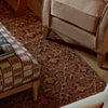 A close-up of a patterned area rug in warm brown, rust, and beige tones, shown in a styled living room setting with part of a beige armchair and a matching ottoman visible.