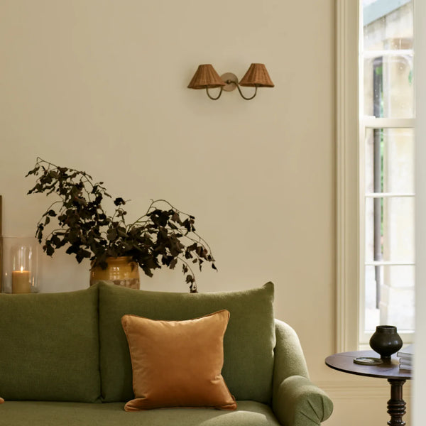 Athena brushed brass double wall light with rattan lampshades on cream wall in living room with olive green sofa, dark wooden side table, large vase and candle in hurricane vase