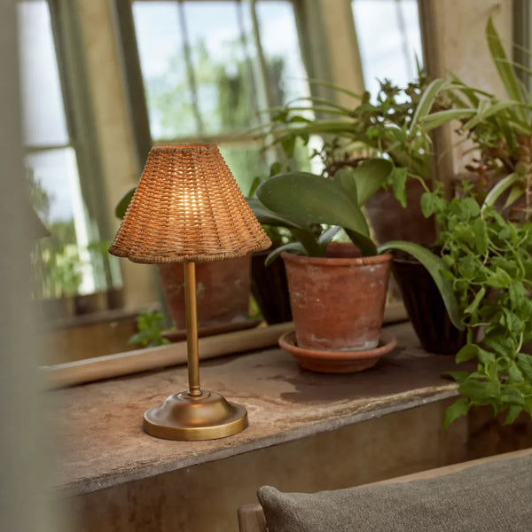 Athena Portable brass and rattan table lamp in garden room next to mirror and plants in terracotta pots, light on