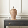 A woven natural rattan table lamp with a ribbed vase shape and a black socket cap, displayed on a dark gray wooden console table against a white paneled wall.