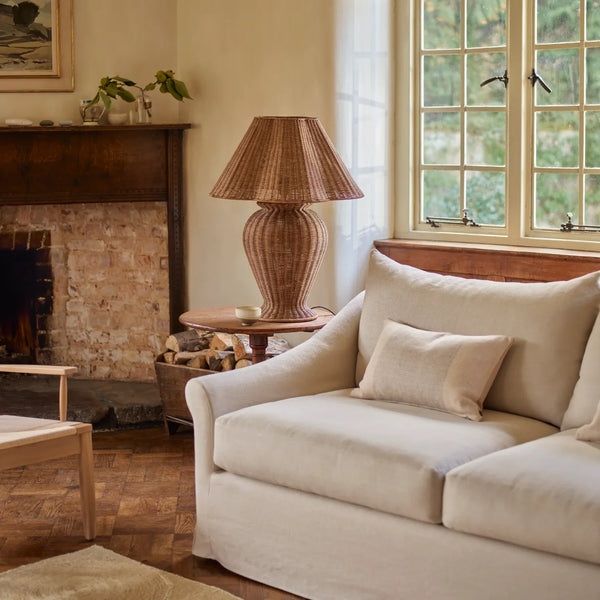 A cream-colored fabric sofa in a warm, rustic living room setting with a brick fireplace, a wicker lamp on a wooden side table, and a large window.