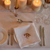 A styled table setting with a white linen napkin featuring a small embroidered floral branch motif in the center, placed on a white plate. The setting includes silver forks, knives, a spoon, clear drinking glasses, and lit cream-colored pillar candles on a beige tablecloth.