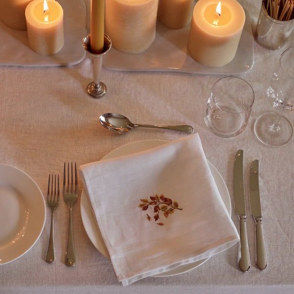 A styled table setting with a white linen napkin featuring a small embroidered floral branch motif in the center, placed on a white plate. The setting includes silver forks, knives, a spoon, clear drinking glasses, and lit cream-colored pillar candles on a beige tablecloth.