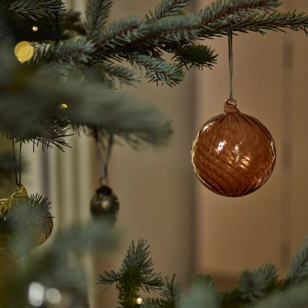 A textured copper-colored glass Christmas ornament hanging from a pine tree branch with warm string lights in the background.