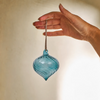 A hand holds a small, teardrop-shaped blue glass ornament with a hanging loop against a warm, softly lit beige background.