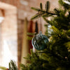 A clear glass Christmas ornament with a swirled blue-green tint hangs from a green Christmas tree branch in front of a softly blurred indoor background.