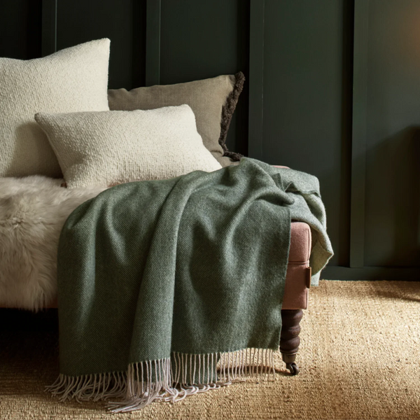 A sage green woven throw blanket with cream fringe is draped over a dusty pink armchair in a styled room setting with dark green paneled walls, neutral pillows, and a textured beige rug.