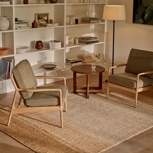 A styled living room scene featuring two mid-century modern armchairs with light wood frames and sage green cushions, arranged on a large natural jute rug around a small round wooden coffee table. In the background are white built-in bookshelves with books and decor, a floor lamp, and a wall art print.