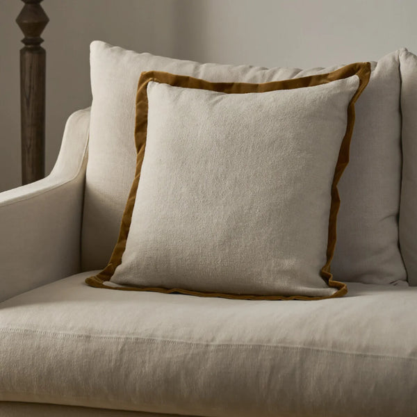 A square throw pillow in a textured cream linen fabric with a contrasting mustard-brown velvet border, styled on a light beige upholstered armchair.