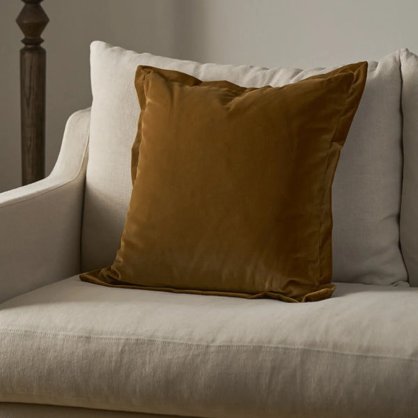 A square throw pillow in a rich camel brown velvet fabric, styled on a light beige linen armchair next to a neutral white pillow.