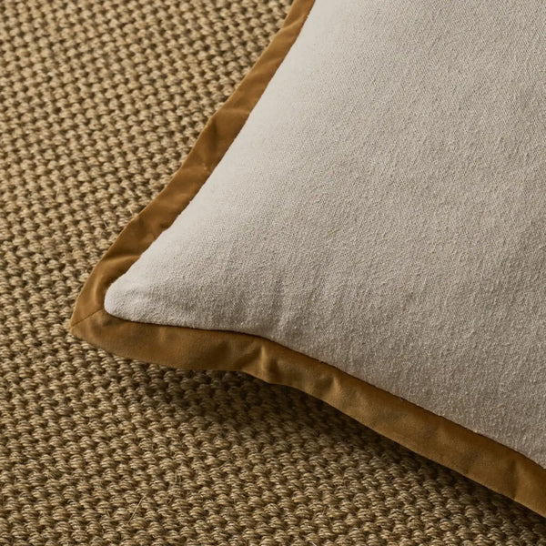 A close-up of a square throw pillow with a light beige textured fabric body and a contrasting mustard brown border, resting on a woven brown rug.