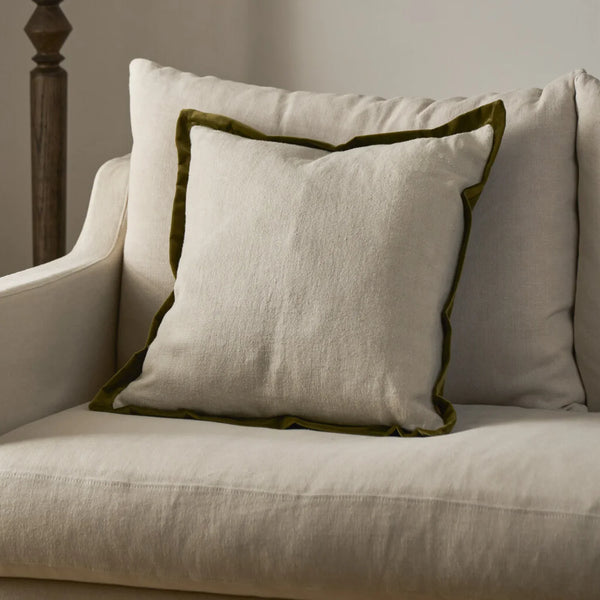 A square off-white linen throw pillow with an olive green velvet piped border, styled on a cream-colored sofa.