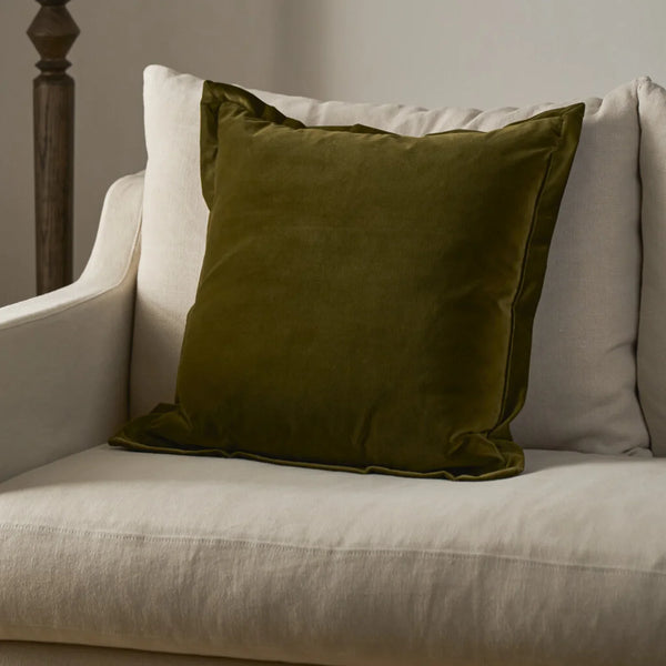 An olive green velvet throw pillow with piped edges resting on a light beige upholstered armchair.