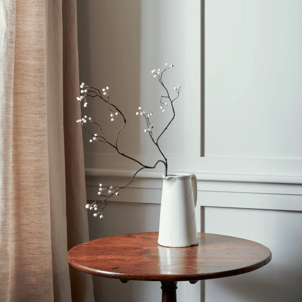 A white ceramic pitcher vase holding dried black branches with small white berries, placed on a round wooden table in a styled interior setting with a beige curtain and white paneled wall.