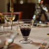 A clear stemmed wine glass with a decorative wavy rim, partially filled with red wine, set on a softly blurred dining table with candles and holiday lights in the background.