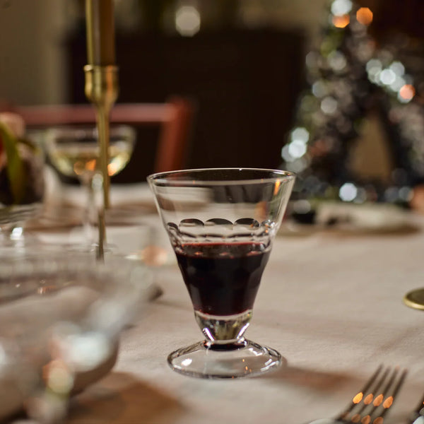 A clear stemmed wine glass with a decorative wavy rim, partially filled with red wine, set on a softly blurred dining table with candles and holiday lights in the background.