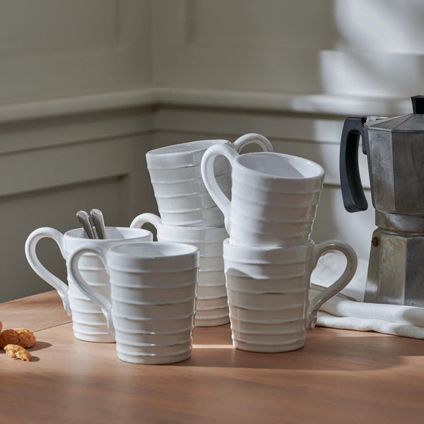 A set of white ceramic mugs with a ribbed texture and curved handles, arranged on a wooden table next to a silver moka pot.
