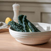 A white ceramic bowl with a ribbed exterior and small beaded trim, filled with dark green curly kale, set on a light wood table with a blurred kitchen background.