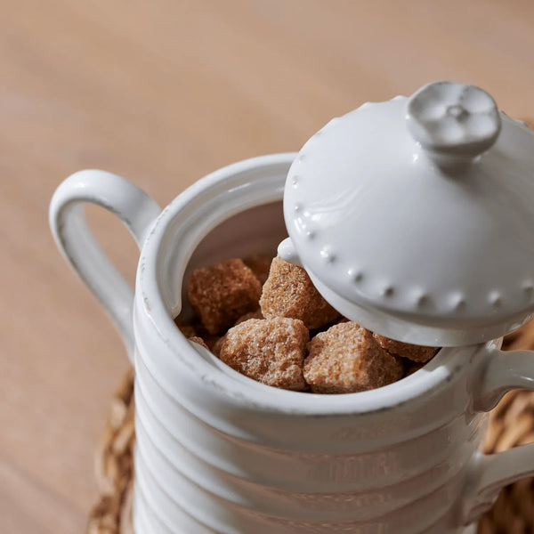 A white ceramic sugar bowl with a matching lid, shown open to reveal brown sugar cubes inside, set on a woven coaster over a light wood surface.