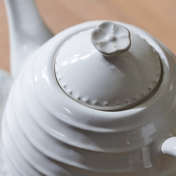 Close-up of a glossy white ceramic teapot showing the lid, floral-shaped handle knob, and ribbed body against a light wooden background.