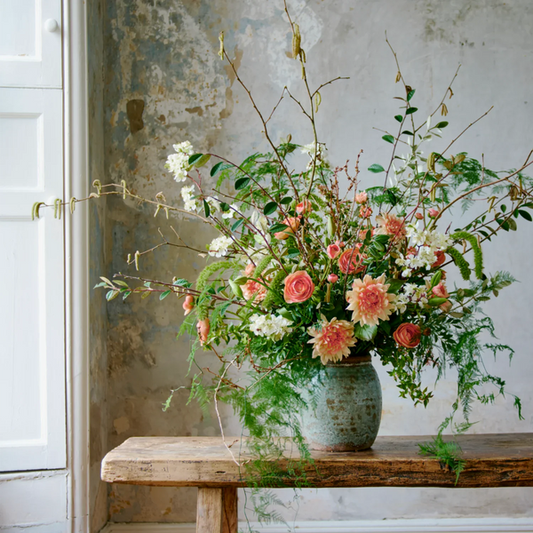 A rustic floral arrangement in a distressed teal ceramic vase, placed on a weathered wooden bench against a textured plaster wall.