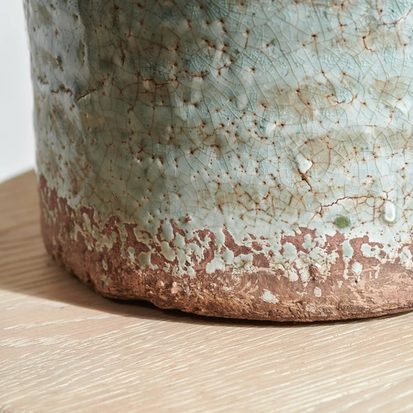 Close-up of a handmade ceramic pot with a textured, crackled glaze in pale blue-green and earthy brown tones, resting on a light wood surface.