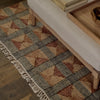 A close-up of a woven area rug with a geometric pattern in beige, brown, and gray, featuring white fringe along one edge. The rug is styled in a home setting under a light-colored upholstered bench on a dark wood floor.