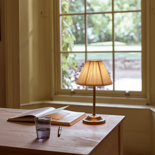Cecile portable brass table lamp with pleated linen shade on a wooden desk in front of window overlooking a garden