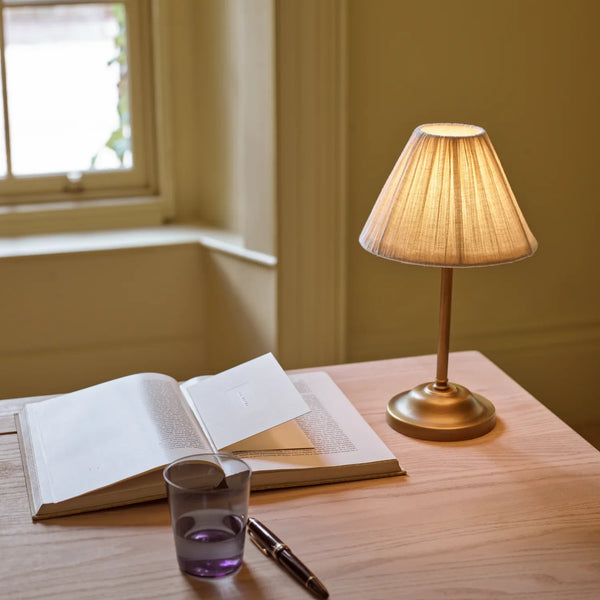 Cecile portable brass table lamp with pleated linen shade on a wooden desk