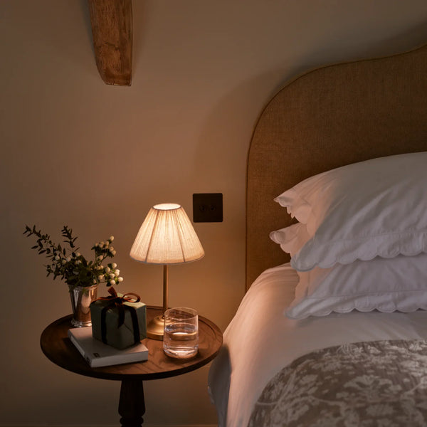 Cecile portable brass table lamp with pleated linen shade on a wooden bedside table in cosy bedroom
