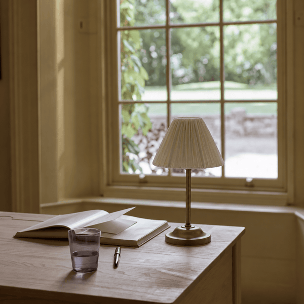 Cecile portable brass table lamp with pleated linen shade on a wooden desk in front of window overlooking a garden