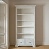 A tall white wooden bookcase with five open shelves and two small drawers at the bottom, set against beige walls and carpeted flooring.