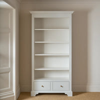 A tall white wooden bookcase with five open shelves and two small drawers at the bottom, set against beige walls and carpeted flooring.
