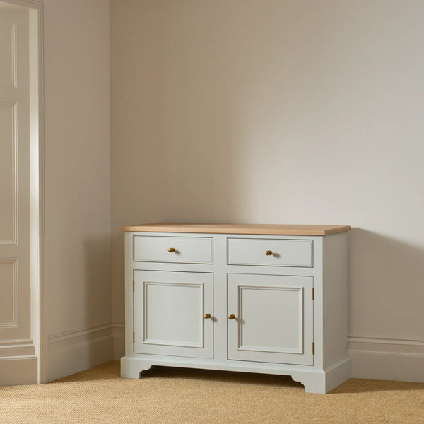 A small white sideboard cabinet with a natural wood top and brass handles, positioned in a corner of a room with beige walls and carpet.