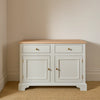 A classic two-door sideboard cabinet with a light natural wood top, painted in a soft off-white/cream finish, featuring two drawers on top and two cabinet doors below with small brass knobs, set against a neutral beige wall and textured beige carpet.