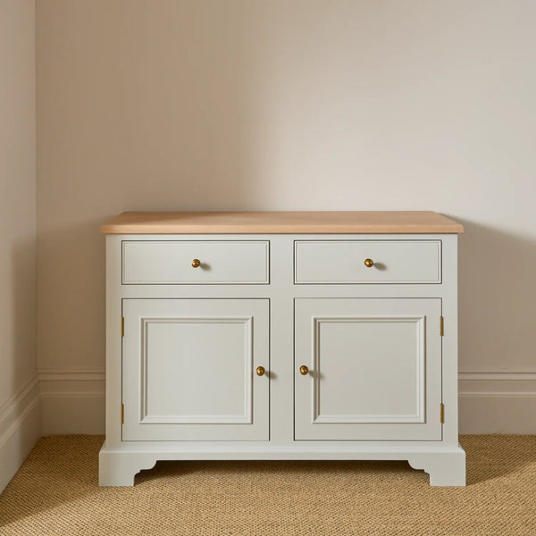A classic two-door sideboard cabinet with a light natural wood top, painted in a soft off-white/cream finish, featuring two drawers on top and two cabinet doors below with small brass knobs, set against a neutral beige wall and textured beige carpet.