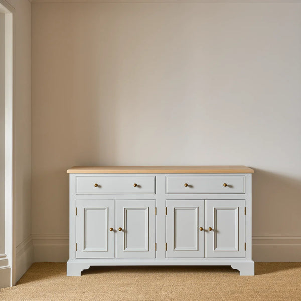 A white wooden sideboard cabinet with a light natural wood top and brass hardware, set against a beige wall and carpeted floor.