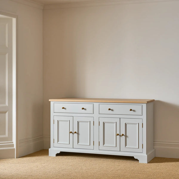 A white wooden sideboard cabinet with a light natural wood top and brass handles, set against a beige wall and carpeted floor.