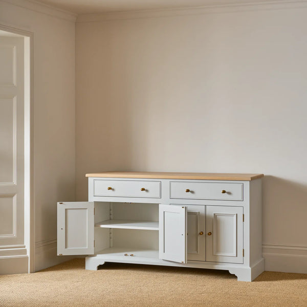 A white wooden sideboard cabinet with a light natural wood top, featuring two drawers on top, two cabinet doors on the bottom with brass knobs, and one open door showing an interior shelf. The cabinet is staged in a room with beige walls and carpet.